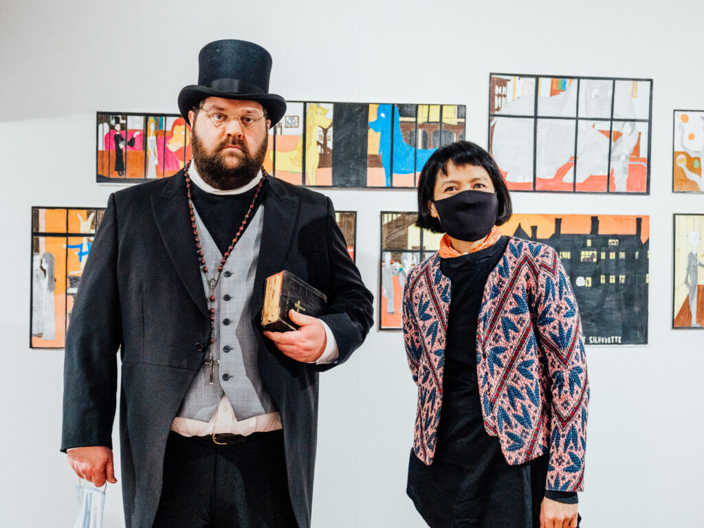 Artists William Gregory, in a Victorian suit, and Mawarini, in a maroon and blue cardigan, stand in front of William’s artworks.