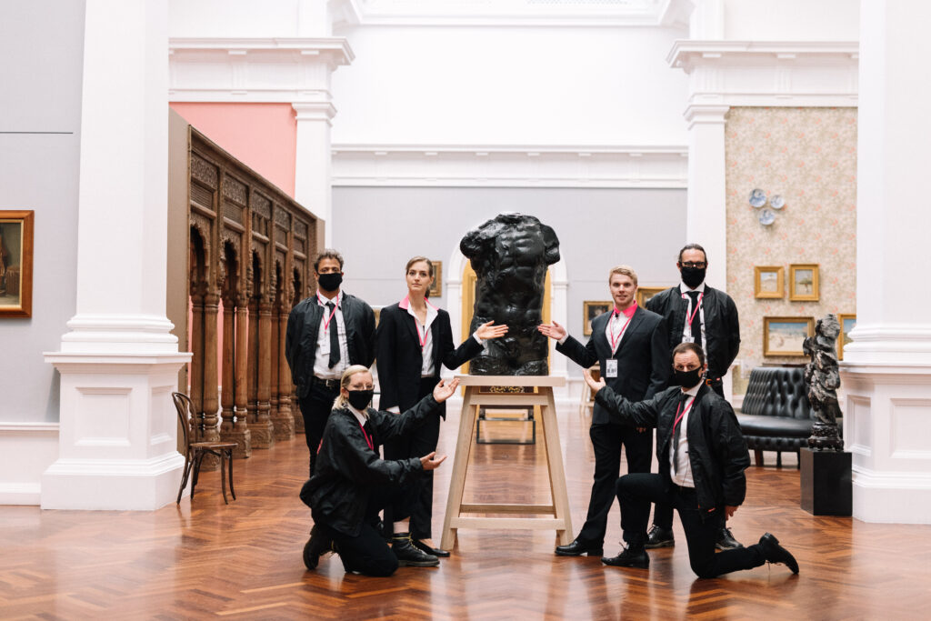 People in suits hold their hands out towards a bronze statue of a torso.