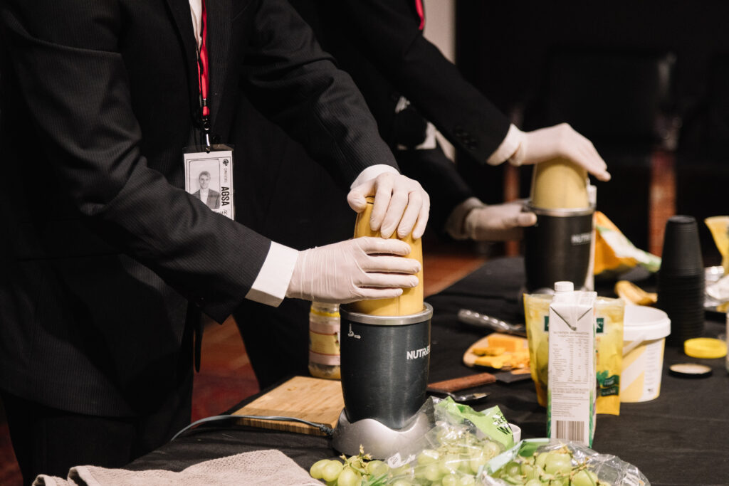 Artists in suits press down on blenders full of a yellow liquid.