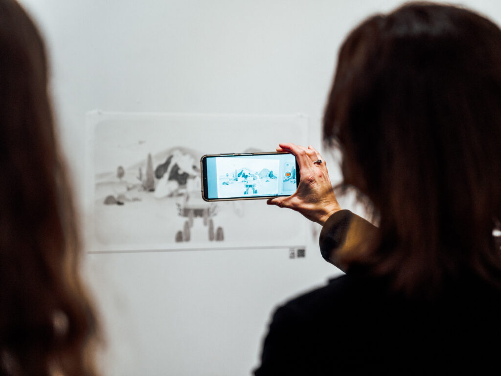 A person holds up their phone, using an app, in front of Mawarini’s artworks.