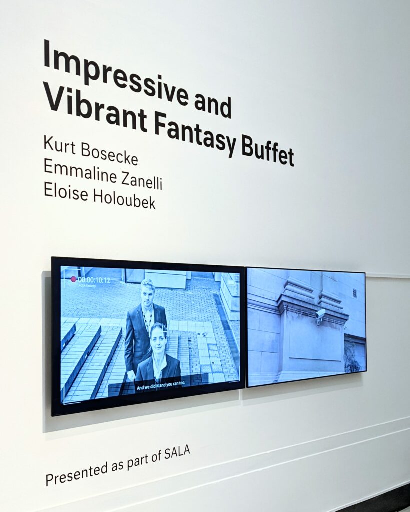 A wall with large black text, Impressive and Vibrant Fantasy Buffet / Kurt Bosecke, Emmaline Zanelli, Eloise Holoubeck, above two wide video screens.