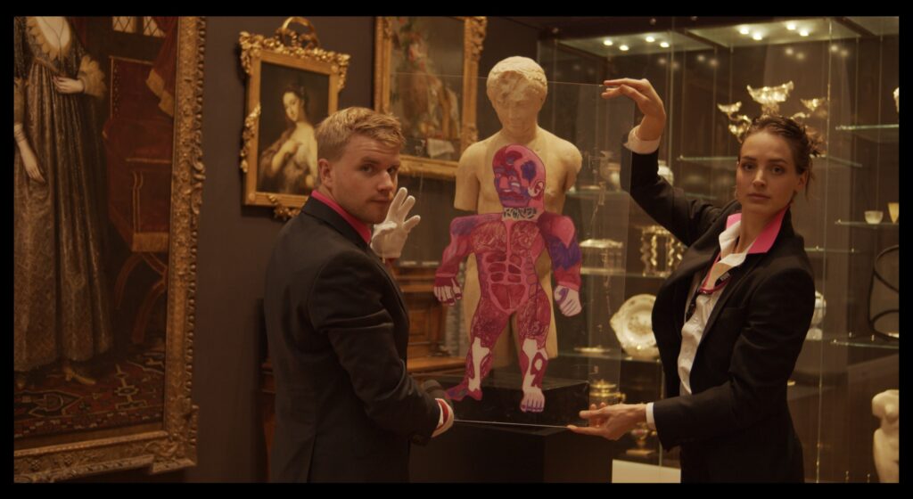 Artists Kurt Bosecke and Emmaline Zanelli hold an anatomical drawing on glass up in front of a marble sculpture of a torso.