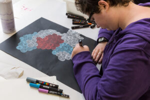 Artist Tori Bedford, a white woman in a purple jumper, drawing at a desk.