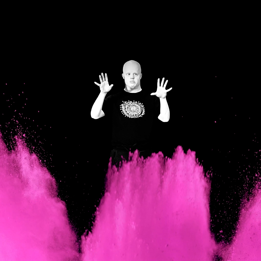 Lorcan Hopper, a white male with a shaved head, stands with his hands up. Pink powder erupts across the bottom half of the image.